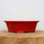 A side profile of a nine-inch glazed ceramic bonsai pot in a vibrant, high-gloss red finish. The pot features an elegant, slightly flared design with a decorative, scalloped or lobed rim and a matching base. It sits on four integrated, subtle feet that slightly elevate the planter. The bonsai pot is displayed on a thick, rustic wooden block, highlighting the contrast between the polished scarlet glaze and the natural wood grain. The entire composition is set against a plain, off-white background, emphasizing the pot's bold color and classic shape.