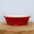 A nine-inch rectangular ceramic bonsai pot with a vibrant, high-gloss red glaze on its exterior and a smooth off-white interior. The pot features an elegant scalloped or lobed design along its flared rim and base, sitting on small, integrated apron-style feet that provide necessary elevation for drainage. The shallow depth and bold primary color make it an ideal planter for shohin or medium-sized bonsai trees, offering a striking aesthetic contrast to green foliage or flowering species. The pot is pictured centered on a rustic, thick-cut circular wooden slab against a clean, neutral white background.