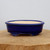 A side-view studio shot of a deep cobalt blue glazed ceramic bonsai pot. The pot has a smooth, oval shape with a low profile and a slightly flared, rounded rim. Its exterior is finished in a high-gloss, dark blue glaze, while the interior is a natural, unglazed beige ceramic. The pot is supported by four short, decorative feet featuring subtle ornamental scrollwork. It is centered on a light-colored, cross-cut wooden base against a plain white background, highlighting its rich color and classic design for bonsai enthusiasts.