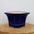 A deep cobalt blue glazed ceramic bonsai pot with an octagonal shape, featuring a flared top rim and decorative recessed panels on each of its sides. The glossy finish highlights the rich, dark blue color. The pot rests on a tiered base with small integrated feet to provide elevation and drainage. The interior of the pot is left unglazed, showing a natural light beige clay body. It is displayed on a thick, circular wooden block with visible grain, set against a plain, neutral white background. This elegant glazed bonsai pot is designed for displaying small to medium-sized bonsai trees.
