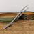 A professional carbon steel bonsai combination tool, featuring a pair of long-reach tweezers and a curved spatula, is displayed on a weathered wooden tabletop. The tool is positioned diagonally across the frame; its broad, flat spatula end rests on the light brown, scratched wood in the lower-left corner, while the long, slender tweezer arms extend upwards toward the top-right. The tweezers feature fine, serrated tips for precision work.

The tool is propped against two shallow, handmade ceramic bonsai pots. The pot on the left has a simple, speckled sage-green and earthy-brown glaze. The pot on the right is more ornate, featuring a deeply textured, carved exterior with a complex glaze of teal, blue, and dark brown. The background is a clean, neutral off-white wall, which makes the metallic grey of the carbon steel tool and the rich textures of the ceramic pots stand out. This image highlights a versatile piece of equipment essential for delicate bonsai maintenance tasks like repotting, weeding, and soil grooming.