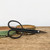 A pair of black carbon steel Tinyroots bonsai shears, part of an ultimate tool kit for bonsai enthusiasts, lies horizontally across a weathered, light brown wooden surface. The professional-grade pruning shears feature slender handles that curve into large, elegant circular finger loops and short, sharp pointed blades held together by a silver-colored pivot screw. In the soft-focus background, two shallow, handcrafted ceramic bonsai pots with a mottled green and earthy brown glaze sit behind the tool. The minimalist composition, set against a plain, off-white wall, highlights the high-quality craftsmanship and aesthetic design of these essential bonsai tools, perfect for precision trimming and maintenance in any starter kit.