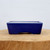 A rectangular, six-inch glazed ceramic bonsai pot in a deep, vibrant cobalt blue color is centered in the frame. The pot features a smooth, glossy finish that reflects light, and its sides slightly taper downward toward the base. At the bottom center, there is a distinct arched cutout that forms two wide feet, a traditional design element for bonsai pots to ensure proper drainage and air circulation for tree roots. 

The top rim of the pot shows a thin, pale beige line where the blue glaze meets the unglazed ceramic body. It is positioned on a rustic, light-toned wooden pedestal that showcases a natural grain and circular texture, likely a cross-section of a log. The background is a minimalist, out-of-focus off-white wall, which makes the rich blue hue of the pot stand out as the primary subject. This type of colorful glazed pot is often chosen for deciduous or flowering bonsai trees to provide a striking visual contrast to the foliage and blooms.