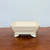 A rectangular 5-inch cream glazed Yixing bonsai pot, model YX2043, sits centered on a rustic, brown wooden surface. The ceramic pot features softly rounded corners and a flat rim, with a subtle crackle pattern visible across its glossy, off-white glazed surface. It is elevated by four sturdy, decoratively carved cloud-style feet. The background is a plain, light-gray wall, which highlights the pot's elegant and simple design, making it a versatile choice for a variety of bonsai tree styles.