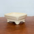 A rectangular, 5-inch cream-glazed Yixing bonsai pot is shown from a slightly elevated side angle. The pot features a glossy, off-white finish with a subtle crackle or crazing pattern throughout the glaze, giving it a classic, vintage appearance. It has an elegant design with slightly flared sides and an inset rectangular panel on the exterior for added texture. The pot is supported by four ornate, outward-curved "cloud" style feet that provide elevation. It sits on a dark brown, polished wooden tabletop against a plain, light-colored wall, highlighting the clean lines and craftsmanship of the ceramic vessel. This type of Yixing pot is a traditional choice for displaying small bonsai trees, offering both aesthetic appeal and practical drainage.