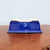 A rectangular glazed bonsai pot in a deep cobalt blue with a high-gloss, reflective finish. The pot is shown upside down on a warm-toned wooden tabletop, revealing its unglazed, cream-colored ceramic base. This base features four sturdy integrated feet at the corners and a central drainage hole, highlighting its practical design for bonsai cultivation. 

The sides of the pot taper slightly inward toward the top, leading to a subtle flared rim resting on the wood. The glossy blue glaze shows rich depth and slight color variations, suggesting a handcrafted quality. The background is a simple, neutral light blue-grey wall, putting the focus entirely on the craftsmanship of the ceramic vessel.