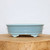 A shallow, oval-shaped light blue glazed bonsai pot is centered on a thick, circular slab of light-colored natural wood. The ceramic pot features a smooth, sky-blue glaze with a subtle sheen and very fine speckling. It has a prominent, rounded upper rim and a decorative horizontal line wrapping around the base. The pot is elevated by four elegant cloud-style feet, two of which are visible in the foreground. The entire scene is set against a plain, off-white background, putting all focus on the pot's classic design and soft color.