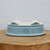 A detailed view of an oval-shaped, light blue glazed bonsai pot shown from a low-angle perspective while resting upside down on a thick, natural wood stump. The ceramic pot features a smooth, glossy exterior in a soft sky-blue hue that beautifully catches the light. Four decorative feet, each intricately carved with a classic cloud or scrollwork motif, are also finished in the same light blue glaze. The unglazed bottom of the pot reveals a creamy, off-white ceramic body with several circular drainage holes distributed across its surface, essential for maintaining proper soil moisture. The minimalist, light-grey background focuses all attention on the elegant design and tranquil color of this professional-grade bonsai container.