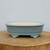 A side-view profile of a shallow, oval light-blue glazed bonsai pot from Bonsai Outlet. The pot features a smooth, light powder-blue glazed exterior with a soft sheen, while the interior remains an unglazed, natural cream-colored clay. Its design includes a gently rounded body, a subtle horizontal band near the bottom, and four decorative feet adorned with traditional cloud-like scroll patterns. The pot sits centered on a thick, cross-cut wooden block against a plain, light grey background, highlighting its elegant shape and craftsmanship.
