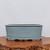 A detailed photograph of a light blue glazed ceramic bonsai pot, showcasing its elegant oval shape and soft, pale blue-gray finish. The pot is designed with a thick, rounded rim and a decorative, scalloped footed base that provides both stability and aesthetic appeal. Its smooth, semi-glossy surface has an even color throughout, reflecting soft light. Resting on a rustic, thick wooden slab with a natural, irregular bark edge, the pot is presented against a clean, neutral white background. This high-quality bonsai container's interior shows a dark reddish-brown hue, typical of traditional stoneware.