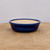 A close-up, eye-level shot shows a shallow, round blue glazed bonsai pot resting on a textured, light brown wooden surface. The pot features a rich, deep cobalt blue exterior with a glossy finish and a thick, rounded lip. The interior is left unglazed, revealing a natural, light beige ceramic material that provides a rustic contrast to the polished exterior.

The pot is centered in the frame against a clean, neutral off-white background, which draws focus to its elegant craftsmanship and simple design. The natural grain and warm tones of the wooden surface beneath add an organic feel, illustrating how the ceramic container would look as a functional and aesthetic base for a bonsai tree. The soft, even lighting highlights the smooth curves and uniform color of the glaze, making it a classic choice for various tree styles.