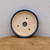 A top-down view into a shallow, round ceramic bonsai pot featuring a deep blue glazed outer rim. The interior of the pot is a light cream or beige color, showing the natural ceramic material. At the center of the base is a large circular drainage hole, with three smaller holes arranged around it for anchoring bonsai wire. The number '101' is hand-written in pinkish-red ink on the inner bottom surface. The pot sits on a textured brown wooden or cork-like surface against a plain, light grey background, highlighting its design and functional features for bonsai growers.