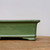 A side-profile, close-up shot of a small, rectangular bonsai pot with a glossy green glaze. The pot features a distinct flared upper rim and sits on short, elegantly curved feet that flare outwards. Its surface reflects soft light, highlighting its smooth, lustrous finish and deep olive-green color. The pot rests on a natural, light-brown wooden surface with a visible grain, set against a simple, neutral-toned white wall that draws focus to the details of the ceramic craftsmanship. This six-inch glazed bonsai container is designed with a classic aesthetic, ideal for pairing with a variety of small bonsai tree species.