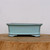 A side-view photograph of a six-inch rectangular glazed bonsai pot in a serene light blue color. The ceramic pot features an elegant flared upper rim and a slightly tapered body that widens again at the base into integrated, low-profile feet. The soft blue glaze has a smooth, glossy finish, accented by delicate dark, antiqued detailing along the edges and corners, giving it an artisanal look. The pot is positioned in the center of a thick, natural-grained wooden block, set against a plain, off-white background that emphasizes its clean lines and soft color palette.