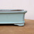 A close-up, detailed side profile of a six-inch light blue glazed ceramic bonsai pot. The pot features a smooth, glossy sky-blue celadon glaze with subtle fine speckling. A dark, brownish-grey distressed finish accents the top rim and the vertical corner, giving it an elegant, aged appearance. The rectangular pot has a slightly flared upper edge and sits on a low, decorative corner foot. The container is placed on a flat, tan wooden surface against a soft, off-white background, highlighting the delicate color and craftsmanship of the glazed pottery.