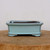An eye-level, front-facing photograph of a six-inch rectangular ceramic bonsai pot featuring a smooth, pale light-blue glaze. The shallow pot has a traditional silhouette with a slightly outward-curving rim and small, decorative feet at each corner. The robin's egg blue finish shows subtle dark antiquing and earthy undertones along the edges and corners, giving the piece a classic, weathered character. The pot is empty, revealing a dark interior, and is centered on a thick, circular wooden block showing natural grain and texture. The background is a clean, neutral off-white, focusing all attention on the craftsmanship and serene color of the glazed bonsai container.