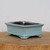 A six-inch rectangular glazed bonsai pot in a soft light blue color, featuring subtle dark brown distressing along its flared top and bottom edges for a vintage, antique look. The ceramic pot has four small, integrated feet at the corners to provide elevation. Its smooth, glossy exterior contrasts with the dark, unglazed clay visible on the interior. The pot is centered on a rustic, circular wooden block against a clean, neutral white background, highlighting its elegant shape and serene color palette.