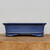 A long side-view shot of an eight-inch cobalt blue glazed ceramic bonsai pot, resting on a rustic, light-colored wooden slab against a neutral off-white background. The rectangular pot features a rich, deep blue glaze with a fine, speckled texture. Its design includes elegantly flared rims and soft, inset corners that give it a refined, slightly indented shape. Near the bottom, subtle horizontal molded lines provide a decorative base, supported by four short, sturdy feet with a curved silhouette. The clean, professional presentation highlights the pot's vibrant color and classic craftsmanship, making it a sophisticated choice for displaying various bonsai tree species.