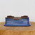 An 8-inch rectangular blue glazed bonsai pot from Bonsai Outlet, featuring a vibrant, mottled cobalt blue glaze with fine speckling across its exterior. The ceramic pot has a distinctive architectural shape with indented corners and a subtly flared base, giving it an elegant, classic profile. The top rim and interior base are unglazed, revealing a dark brown natural clay body, while several drainage holes are visible on the inside to ensure proper soil aeration and water management for a bonsai tree. The pot sits centered on a rustic, thick circular light-wood block against a clean, minimalist off-white background, highlighting its color and craftsmanship.