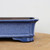 A detailed close-up side view of a corner of an 8-inch rectangular glazed bonsai pot. The pot is coated in a vibrant, mottled blue glaze featuring a fine, salt-and-pepper texture with darker blue and lighter flecks throughout. The design showcases a slightly flared upper rim and a decorative horizontal ridge running along the bottom edge. One of the pot's sculpted, cloud-style feet is clearly visible at the corner, providing a traditional aesthetic. The high-fired ceramic pot rests on a light-colored wooden surface against a clean, out-of-focus white background, highlighting its elegant form and glossy finish.