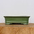 A detailed side-profile photograph shows an eight-inch rectangular green glazed ceramic bonsai pot. The pot features a rich, glossy olive-green finish that reflects light subtly. Its design includes a slightly flared top rim and a decorative horizontal ridge wrapping around the lower portion of the body. The pot is supported by four short, integrated feet, providing space for drainage beneath. It is displayed on a thick, rustic wooden slab with a visible natural grain, set against a minimalist, off-white background, highlighting the pot's classic aesthetic for bonsai enthusiasts.