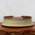A detailed image of a 16-inch cream-colored glazed ceramic bonsai pot, presented on a rustic, dark wood slab with a natural, textured edge against a plain white background. The oval-shaped pot features a distinctive flared base and two horizontal recessed lines circling its exterior for added detail. The exterior glaze is a light beige or cream with a subtle, fine-grained texture, while the top rim is accented with a contrasting dark brown or burnt sienna glaze that blends into the body. Two symmetrical cut-outs on the upper rim provide a unique silhouette. The interior of the pot reveals a smooth, reddish-brown unglazed clay surface with visible drainage holes at the bottom, making it a functional and aesthetic choice for large bonsai trees.
