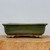 A professional side-profile product photograph of a ten-inch rectangular bonsai pot featuring a rich, mottled moss-green glaze. The glazed ceramic planter has a thick, slightly flared rim and rounded corners, giving it a soft yet sturdy appearance. The pot is elevated on four decorative scroll-style feet, with two visible in the image, each adorned with a subtle carved swirl design. It is displayed on a light-colored, natural wood cross-section base against a clean, neutral light gray background, which highlights the earthy tones and textured finish of the glaze. This elegant green glazed bonsai pot is designed for displaying medium-sized bonsai trees and provides a classic, natural aesthetic.