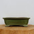 A side-view studio photograph of a nine-inch rectangular bonsai pot with a glossy green glaze, showcasing its classic design. The ceramic planter features a prominent recessed rectangular panel on its front side, a wide, slightly flared rim, and four elegant scroll-style feet at the corners. The glaze is a deep olive green with subtle tonal variations, giving it a handcrafted, mottled appearance. The pot is displayed on a thick, natural cross-section of a wooden log, highlighting its earthy aesthetic against a clean, neutral, off-white background. This glazed bonsai pot is designed for displaying medium-sized bonsai trees, offering a traditional and sturdy base for horticultural art.