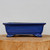 A detailed studio shot of a nine-inch glazed ceramic bonsai pot in a striking, deep cobalt blue. This rectangular planter features a slightly flared upper rim and a long, recessed decorative panel on its front face. The pot is elevated by four ornate, scroll-styled "cloud" feet at each corner, adding a touch of traditional craftsmanship. The vibrant blue glaze has a smooth, glossy, and reflective finish that provides a beautiful contrast. It is displayed resting on a rustic, circular cross-section of a wooden log against a clean, neutral white background, highlighting its elegant design and rich color.
