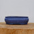 A side-view photograph of a six-inch blue glazed ceramic bonsai pot. The rectangular planter features elegant lobed corners and a slightly flared profile, giving it a classic and sturdy appearance. The glaze is a deep, mottled indigo blue with a fine, speckled texture that adds depth and a stone-like quality to the finish. The pot is designed with a distinct rim and sits on small, integrated feet that provide necessary elevation for proper drainage and air circulation, which are essential for healthy bonsai growth. It is showcased on a thick, rustic slab of light-colored wood against a clean, neutral off-white background, highlighting the pot's artisanal craftsmanship and functional design.