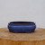 A six-inch glazed blue bonsai pot with a rectangular, lobed shape sits centered on a rustic, circular wooden slab. The planter features a vibrant, mottled blue glaze with fine speckling and subtle variations in shade, giving the surface a rich, textured appearance. Its design includes a prominent rim and a bulbous mid-section that curves inward toward a base supported by four small, integrated feet. The interior of the pot reveals the warm, natural reddish-brown color of the unglazed clay. Set against a clean, neutral off-white background, the deep blue of the ceramic container stands out as an elegant and functional piece for housing a small bonsai tree.
