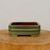 A detailed studio photograph showcases a small, rectangular bonsai pot with a glazed green finish, positioned centrally on a thick, natural wood pedestal with a visible grain. The planter’s exterior is coated in a soft, olive-green glaze that features a fine-textured, semi-matte appearance. Its design is characterized by horizontal raised bands that wrap around the circumference, intersected by subtle vertical lines at the corners to create a classically structured look. The pot rests on four low, integrated block feet, providing a slight elevation. The interior of the pot reveals a contrasting reddish-brown ceramic material. Set against a clean, off-white background, this six-inch green planter is a classic and sturdy vessel designed for the artistic display and healthy growth of miniature bonsai trees.