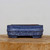 A detailed side view of a six-inch rectangular blue bonsai pot with a speckled glaze finish. The ceramic planter features a unique mottled pattern of deep blue and light, stippled white highlights. A prominent horizontal raised band wraps around the middle of the pot, adding architectural interest to its rounded rectangular shape. The pot sits on four integrated block feet, providing essential elevation for drainage. It is presented on a thick, natural wood pedestal with visible grain, set against a clean, neutral white background to emphasize the rich texture and color of the glazed ceramic.
