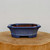 An eye-level shot features a seven-inch glazed ceramic bonsai pot in a vibrant, mottled blue color. The planter has an elegant octagonal shape with sides that flare slightly outward toward a thick, flat rim. The blue glaze has a finely speckled, stone-like texture, while the interior of the pot reveals the unglazed reddish-brown clay beneath the rim. This shallow bonsai planter rests on small, integrated blocky feet, designed to provide stability and essential drainage for a bonsai tree. It is displayed on a thick, natural-grained wooden pedestal against a clean, off-white background, highlighting its artisanal quality.