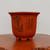 A tall, square-shaped unglazed bonsai pot made of reddish-brown clay, featuring a wide flared rim and four small, sturdy block feet. The front of the pot is decorated with a traditional black ink-style landscape illustration showing stylized mountains, a weeping tree, and a small hut. To the right of the artwork, vertical columns of elegant Chinese calligraphy are etched into the surface. A pattern of small circular aeration holes is visible near the base on one side. The pot sits on a polished, warm-toned wooden surface with a natural, irregular edge, all set against a neutral, plain background. This detailed etched planter exemplifies the classic aesthetic of professional-grade unglazed bonsai pottery.