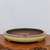 A shallow, 17-inch round glazed ceramic bonsai pot sits centered on a dark, rustic wood slab with a rough, natural bark edge. The pot features a smooth, light-beige or cream-colored glaze on its exterior, which transitions to a dark reddish-brown clay finish on the interior. A delicate decorative band encircles the upper portion of the pot just below the rim, consisting of a thin dark line accented with small, evenly spaced raised studs or rivets. Two drainage holes are visible at the bottom of the pot's interior. The pot is set against a clean, neutral white background, emphasizing its wide, low profile and elegant craftsmanship.