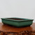 A side-angle, eye-level photograph of a shallow rectangular glazed bonsai pot. The planter is finished in a vibrant, glossy sea-green or teal glaze that reflects light smoothly. It features rounded corners and a slightly flared top edge. The interior and the low-profile base are a contrasting matte, unglazed dark brown ceramic. The pot is displayed on a polished, dark-stained wooden stand with a rustic, irregular natural edge. The background is a plain, light gray wall, focusing all attention on the elegant green ceramic planter.