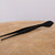 A detailed side-profile view of Kaneshin #58 Arrow Pine Needle Tweezers, a specialized Japanese bonsai tool, resting on a rustic, light-colored wooden surface. The tweezers feature a sophisticated matte black finish. The tool is designed with dual functionality: one end tapers to fine, precise tips perfect for plucking pine needles or delicate pruning, while the opposite end flares into a flat, smooth, arrow-shaped spatula used for tamping soil or weeding. The handles include a textured, ridged section for a secure grip during intricate bonsai maintenance. The background is a clean, neutral white, emphasizing the sleek lines and professional quality of the tool.