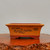 An eye-level, straight-on shot of a rectangular, unglazed ceramic bonsai pot featuring a detailed landscape etching on its front side. The pot exhibits a two-tone color palette with a light, sandy-orange body and a contrasting deeper reddish-brown rim and base. 

The etched artwork depicts a traditional, serene scene with rolling mountains, clusters of stylized trees, a small village with traditional-style houses, and a flock of birds in flight. Small Chinese characters are delicately etched in the upper right corner of the landscape. The pot has a classic rectangular shape with elegantly flared upper edges and is supported by four small, integrated feet. It sits upon a polished, dark wood stand with a rustic, natural-edge finish, all set against a clean and neutral off-white background. This traditional vessel is designed for bonsai trees, blending functional gardening with artistic craftsmanship.