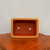 An overhead, slightly angled view of a rectangular unglazed bonsai pot with a classic terracotta-colored finish. The pot features distinctively indented corners, giving it a clean and architectural look. Its thick, sturdy walls have a smooth, matte texture in a light reddish-brown hue. Inside the basin, which has a slightly deeper reddish-orange tone, two large circular drainage holes are clearly visible at the bottom, essential for proper soil aeration and moisture control. The pot rests on a polished wooden stand with a rich, dark amber grain and a natural, uneven edge. The entire composition is set against a minimalist, neutral off-white background, emphasizing the simple and functional design of this ceramic bonsai container.
