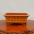 A rectangular, unglazed bonsai pot featuring a two-tone design with a light ochre clay body and a contrasting dark reddish-brown rim and stepped base. The front side of the pot is adorned with two rows of hand-etched Chinese calligraphy characters in black ink. The pot is shown from a straight-on perspective, resting on a highly polished, dark wood stand with a natural, burl-like edge, set against a plain, off-white background.