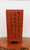 A tall, rectangular unglazed red clay bonsai pot, designed for a cascade-style tree, is captured in a front-facing studio shot. The pot's earthy, terracotta-colored surface features intricate black-etched Chinese calligraphy, with characters flowing down in three vertical columns. This "Etched Cascade" pot has a clean, minimalist silhouette that tapers slightly towards its base, which is elevated by four small, subtly curved feet. The vessel is presented on a polished, warm-toned burl wood stand against a stark, neutral white background, emphasizing the contrast between the dark etchings and the natural texture of the unglazed clay.