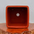 A top-down view looking into the interior of a square, unglazed terracotta ceramic bonsai pot with rounded corners. The pot is a rich, earthy reddish-orange color with a smooth, matte finish. Centered in the bottom is a large circular drainage hole, which is flanked by four smaller holes near each corner intended for wiring a bonsai tree into place. The pot is resting on a highly polished, warm-toned wooden slab with a textured, natural live edge, all set against a neutral off-white background.