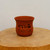 An unglazed, reddish-brown terracotta bonsai pot with a flared rim and rounded base sits centrally on a thick, light-toned natural wood slice. The pot is decorated with traditional black Chinese calligraphy etched onto its side, with large characters across the top and smaller text below. The smooth, matte texture of the clay is highlighted by soft, even lighting, which also showcases the natural grain of the wooden pedestal. Set against a plain, off-white background, the image captures a minimalist and elegant display piece, suitable as a decorative accent or a container for a small bonsai tree.