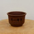 A detailed, eye-level photograph shows a round, unglazed ceramic bonsai pot crafted from smooth, reddish-brown clay. The pot features a matte finish with a wide, slightly flared rim and a body that tapers gently toward a narrow base. 

Centrally featured on the front of the pot is elegant, hand-etched Chinese calligraphy in black ink. The four large characters, "吉祥如意" (Jíxiáng rúyì), are written in a traditional style and translate to "good fortune and as one wishes." Beneath these prominent characters are several columns of smaller, finely etched inscriptions. 

The pot is resting on a light-colored, rustic cross-section of a wooden log, showcasing the natural grain and a small crack in the wood. The background is a clean, neutral off-white, which emphasizes the traditional aesthetic and earthy tones of the etched bonsai container.
