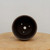 A top-down view looks directly into the interior of a round, unglazed dark brown ceramic bonsai pot. The pot's smooth, matte-finished clay features a large central drainage hole surrounded by three smaller secondary holes designed for bonsai training wires. The pot rests on a light-colored, circular wooden base against a plain off-white background, highlighting the essential functional features for proper bonsai tree health and styling.