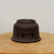 A high-resolution image of an unglazed, dark brown ceramic bonsai pot from Bonsai Outlet, resting upside down on a light-colored wooden surface. The pot, made of smooth, matte clay in a deep chocolate hue, features a graceful, inverted bell shape. Intricately etched onto its side is elegant Chinese calligraphy, with four large, expressive characters set below two rows of smaller, neatly inscribed text. The circular foot of the pot is visible at the top of the image, showing three small, precise cutouts designed for drainage. The entire piece is set against a minimalist, off-white background, highlighting the fine craftsmanship and traditional aesthetic of this etched unglazed bonsai pot.