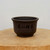 A dark brown, unglazed ceramic bonsai pot sits centered on a thick, light-colored natural wood slab. The pot features a classic rounded, bowl-like shape with a subtle flared rim and a sturdy, slightly raised footed base. Its smooth, matte surface is adorned with traditional Chinese calligraphy etched into the clay and filled with black ink. 

The four prominent characters in the center are written in a fluid, artistic script, translating to "Jíxiáng rúyì," a classic blessing for good fortune and may all things go according to one's wishes. Below this main inscription are smaller vertical columns of more delicate characters, which list various philosophical virtues such as sincerity and professional excellence. The pot’s earthy, dark chocolate tone provides a stark contrast to the pale, grainy wood of the stand and the clean, neutral light-gray background. This traditional piece of pottery exemplifies the understated elegance and cultural artistry often sought in bonsai cultivation.