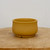 A small, round, unglazed ceramic bonsai pot in a matte mustard yellow or ochre color sits centered on a rustic, light-colored wooden cross-section. The pot features a smooth, bulbous body that tapers slightly toward the top rim and a raised circular base. A small notch for drainage is visible at the bottom edge of the pot's foot. The container is set against a clean, minimalist off-white background, emphasizing the warm, earthy tones of the unglazed clay and the natural grain of the wooden surface.