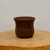 An unglazed bonsai pot in a rich reddish-brown clay, sitting on a rustic, light-colored natural wood pedestal. The pot features a rounded, tapered shape with a subtle flare at the top rim. Etched into the side of the pot is traditional Chinese calligraphy in black ink, including four large, prominent characters and a smaller inscription beneath them. The scene is set against a clean, neutral off-white background, highlighting the earthy tones of the pottery and the organic texture of the wood grain on which it rests.